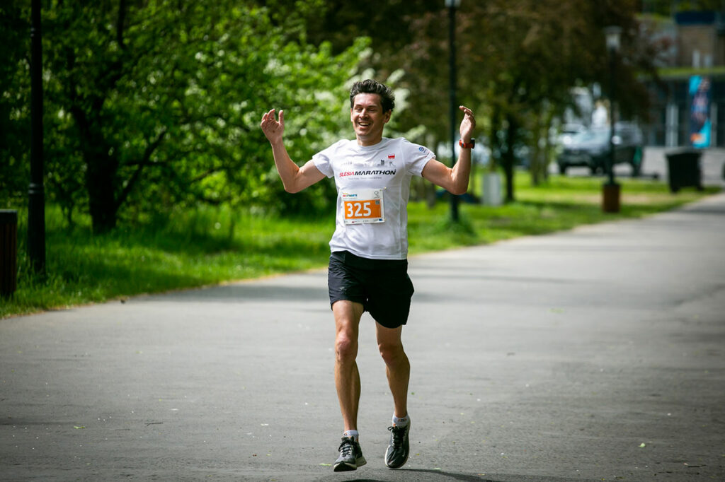 Na tle alejki, wśród drzew, widoczny jest doświadczony biegacz w białej koszulce Silesia Marathon. Na niej ma plakietkę z numerem startowymi i napisem: Dni Otwarte Funduszy Europejskich. Unosi się nad ziemią, podnosi w górę ręce w geście radości i szeroko się uśmiecha.