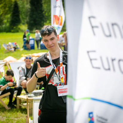 Ilustracją tekstu jest fotografia przedstawiająca młodego chłopaka z okularami przeciwsłonecznymi na głowie i plakietką „wolontariusz” zawieszoną na szyi, który pokazuje gest uniesionego w górę kciuka. Za nim znajdują się uczestnicy pikniku. Fotografia pochodzi z archiwum Urzędu Marszałkowskiego Województwa Śląskiego.