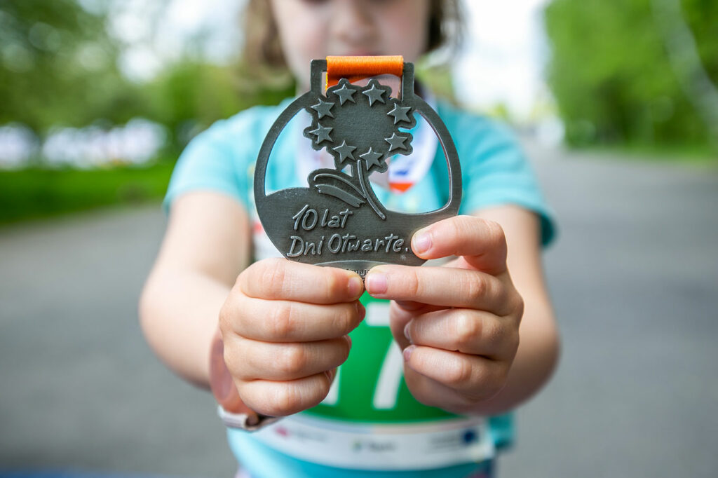Zdjęcie przedstawia dziecko, które wyciąga przed siebie ręce, trzymające pamiątkowy medal z napisem: „10 lat Dni Otwarte”. Fotografia pochodzą z archiwum Urzędu Marszałkowskiego Województwa Śląskiego.