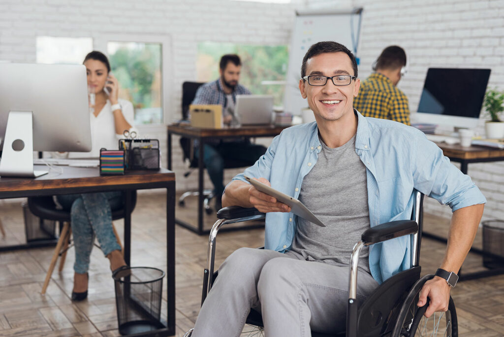 Fotografia ukazuje młodego, uśmiechniętego mężczyznę na wózku inwalidzkim, który znajduje się w biurze. W prawej ręce trzyma tablet.