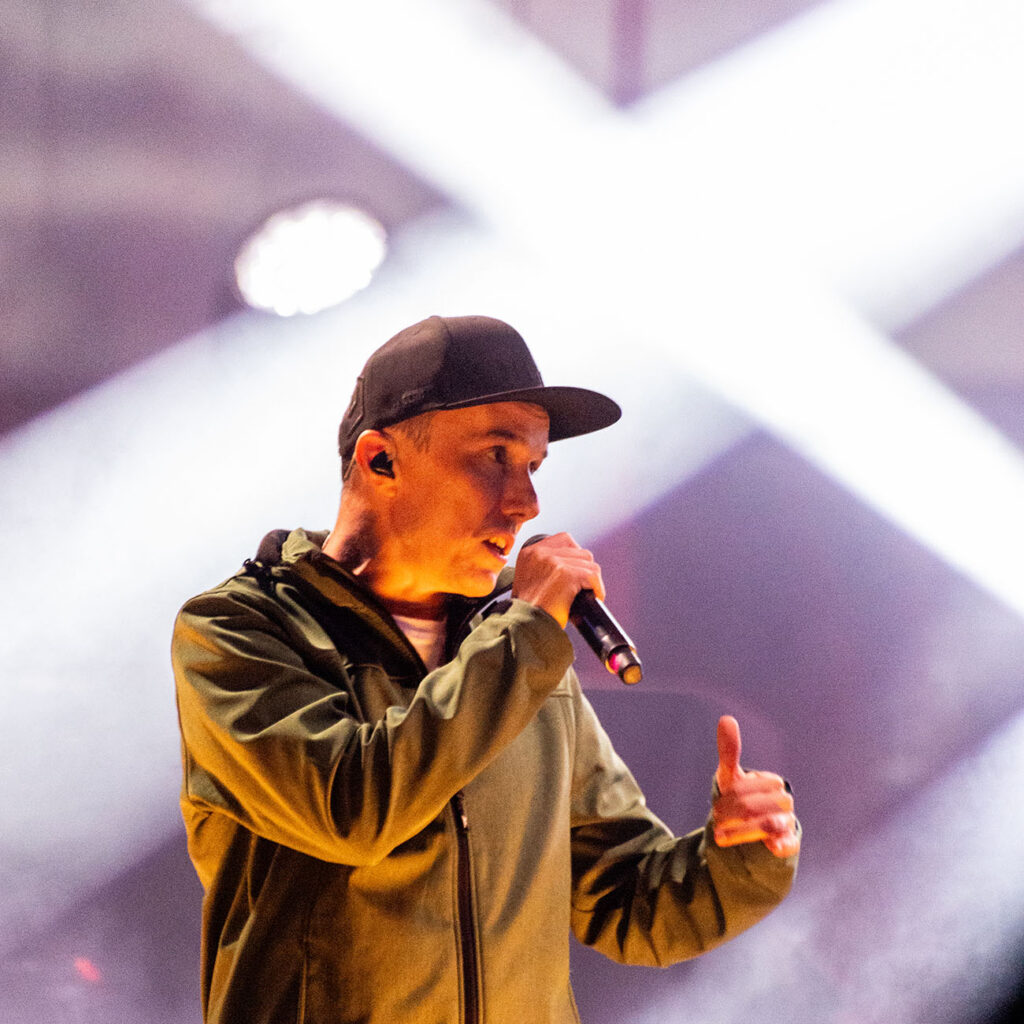 Na fotografii widoczny jest mężczyzna w czapce z daszkiem, który w trakcie koncertu śpiewa na scenie do mikrofonu.