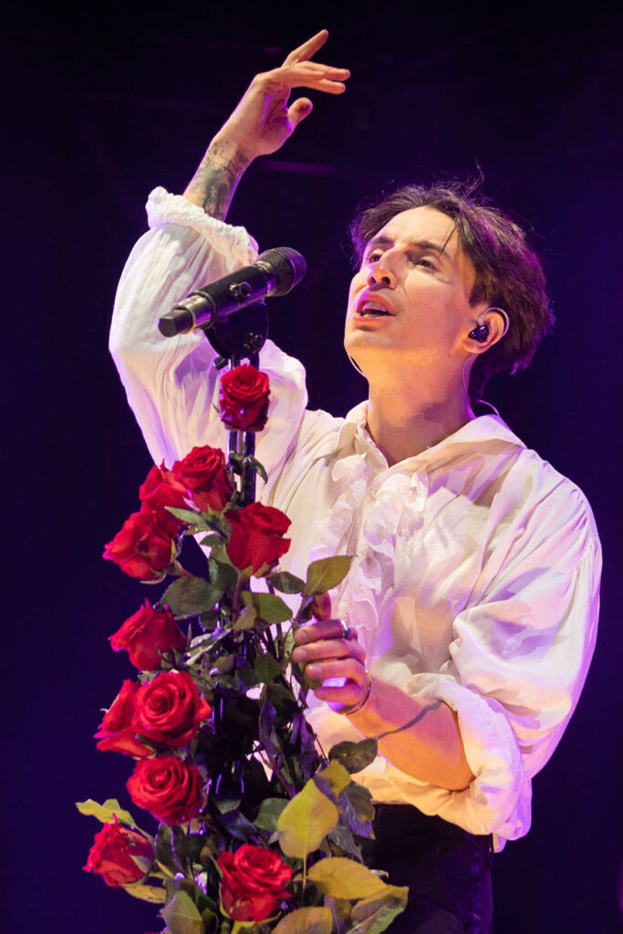 Fotografia przedstawia młodego mężczyznę w białej koszuli śpiewającego na scenie do mikrofonu przystrojonego różami. Ilustracja pochodzi z archiwum Urzędu Marszałkowskiego Województwa Śląskiego.