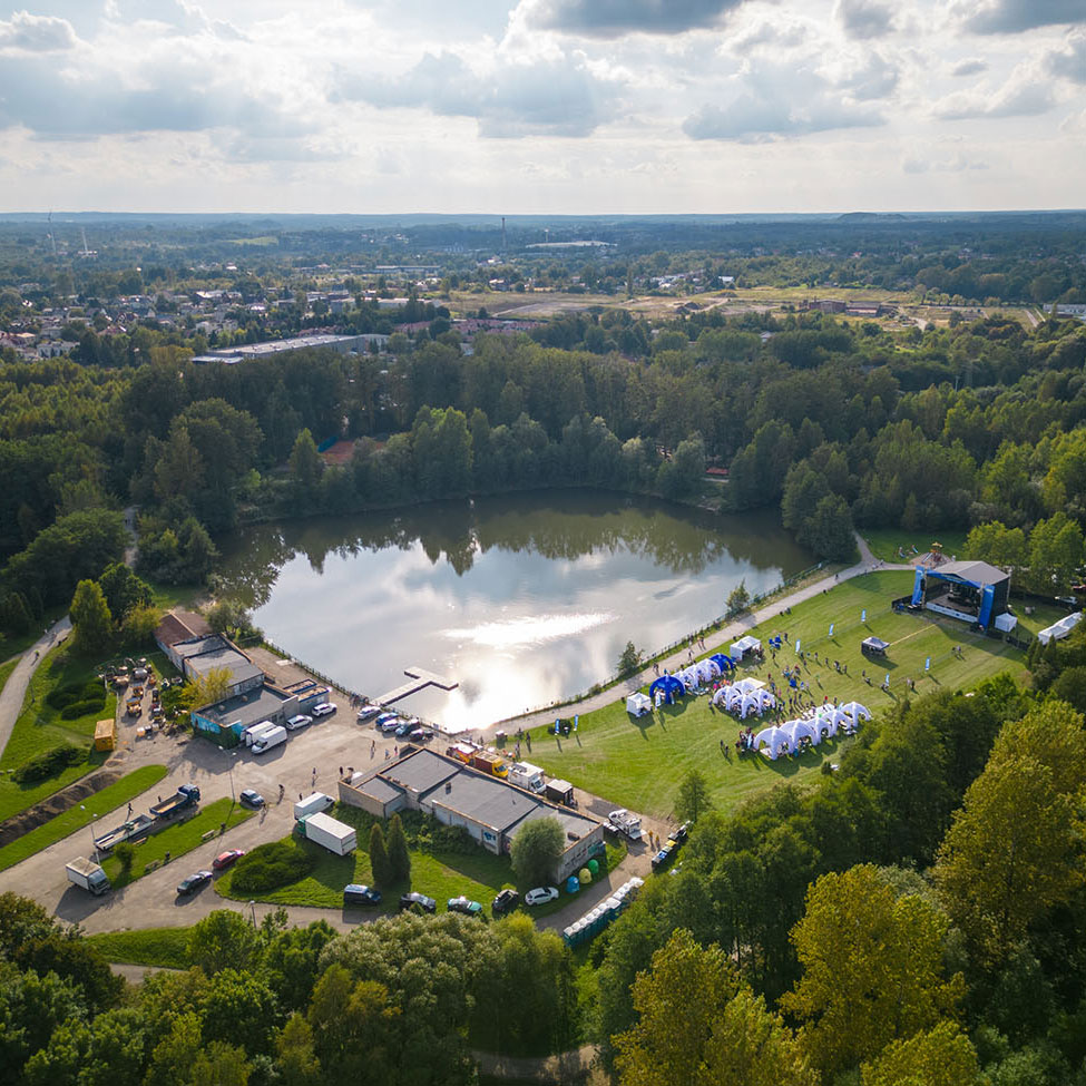 Ilustracja przedstawia park widziany z drona. Na środku jest duży staw, wokół którego znajduje się infrastruktura: budynki i namioty.