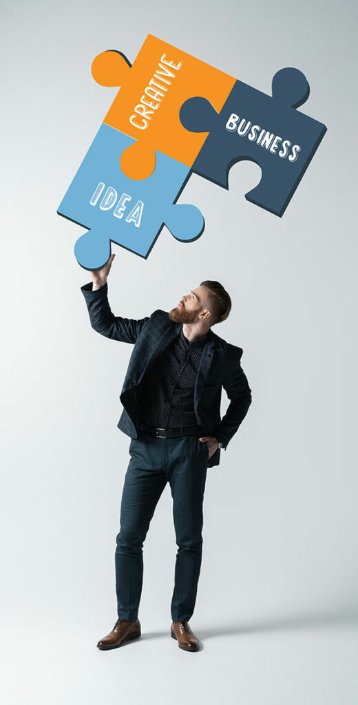 Na zdjęciu jest elegancko ubrany młody mężczyzna, trzymający nad sobą, w prawej ręce, trzy połączone ze sobą puzzle, z napisami: Idea, Creative, Business.