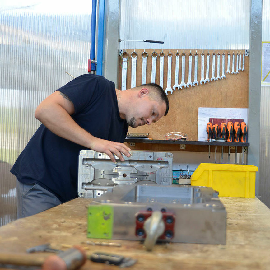 Zdjęcie przedstawia mężczyznę w czasie pracy w warsztacie. Za nim znajduje się panel, na którym zawieszone są śrubokręty i klucze.