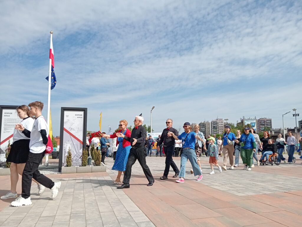 Na zdjęciu przedstawione są osoby na placu, trzymające się za ręce, co wskazuje na taniec poloneza. 