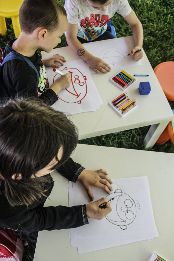 Na zdjęciu widać trójkę dzieci siedzących przy białym stoliku na trawie, zajmujących się rysowaniem. Każde z dzieci rysuje na kartkach papieru, a na stole leżą kolorowe kredki i gumka do mazania. Dzieci rysują różne postacie, a jedna z kartek przedstawia uśmiechniętą twarz. Na pierwszym planie widoczny jest chłopiec w czarnej koszulce rysujący czerwonym flamastrem, a obok niego dziecko w koszulce z postacią Scooby-Doo rysujące czarną kredką. 