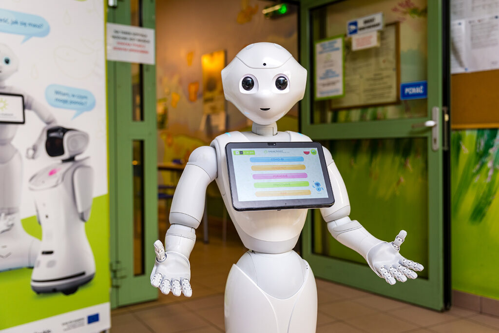 Na zdjęciu widzimy białego robota, który rozkłada ręce. Widoczna jest wyraźnie jego twarz, z oczami i ustami. Na wysokości klatki piersiowej ma zamontowany monitor wyświetlający komunikaty w kolorowych ramkach.