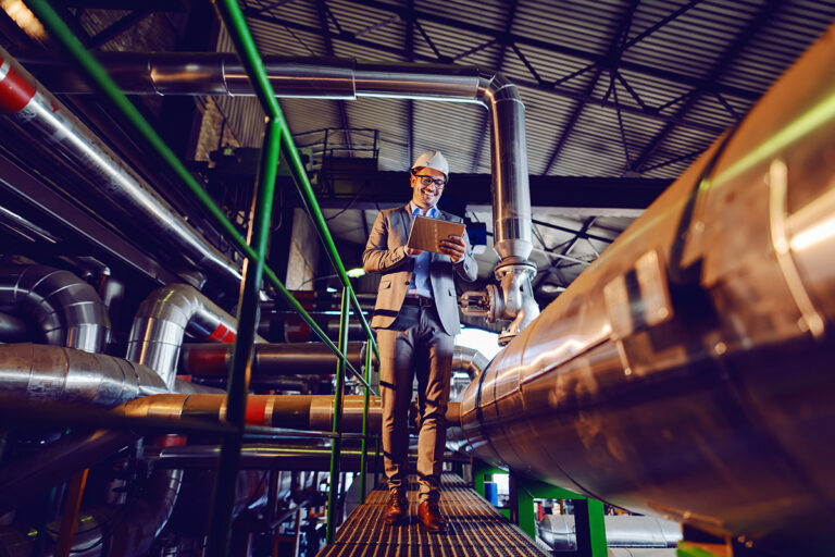 Fotografia przedstawia uśmiechniętego mężczyznę w garniturze i ochronnym kasku, który porusza się stalową kładką biegnącą pomiędzy rurami i zapisuje coś na kartce.