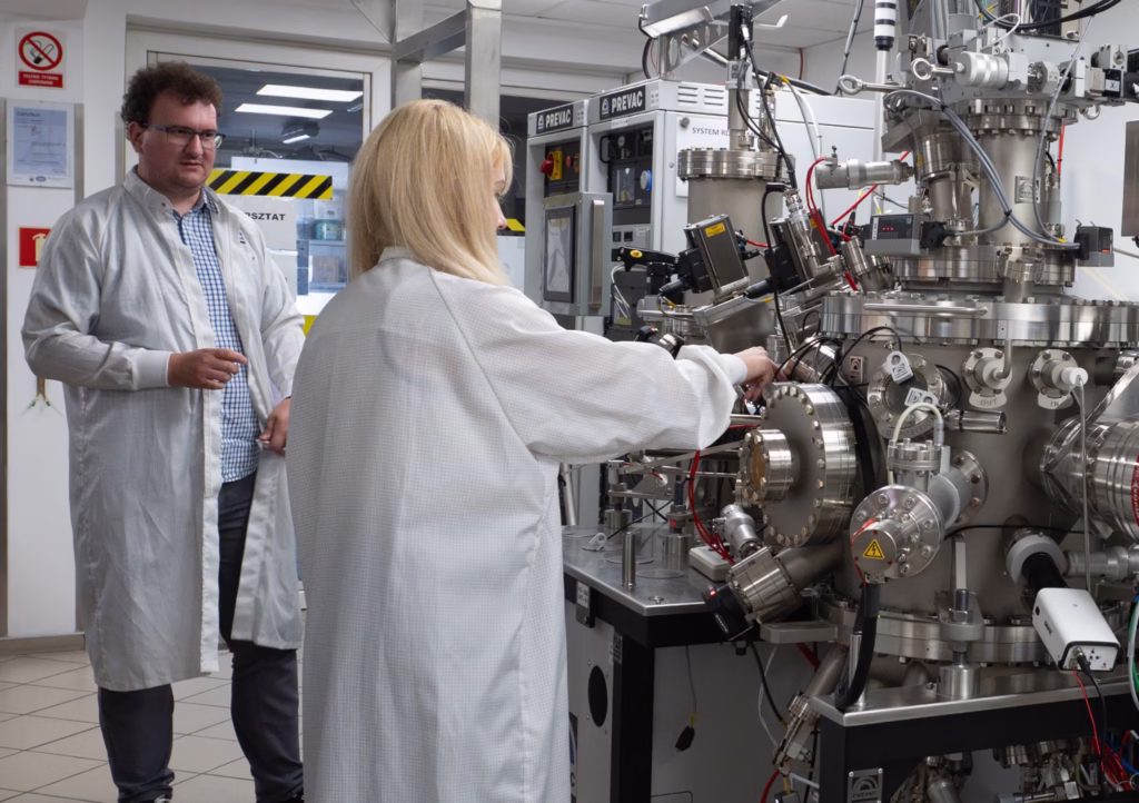 Naukowiec w białym kitlu pracuje przy skomplikowanej aparaturze naukowej, otoczonej różnymi elementami metalowymi, rurami i przewodami. Drugi naukowiec stoi obok, obserwując pracę. Na ścianie w tle widnieją znaki ostrzegawcze oraz inne oznaczenia laboratoryjne.