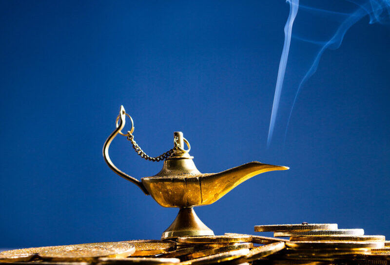 Ilustracja przedstawia Lampę Alladyna, z której wydobywa się dymek. Lampa stoi na monetach.