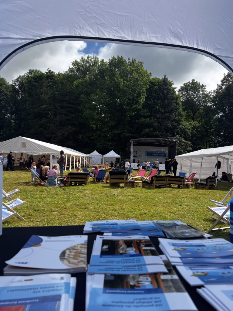 Zdjęcie przedstawia widok na piknik rodzinny z perspektywy stoiska informacyjnego. Na pierwszym planie widać stolik z rozłożonymi ulotkami i broszurami informacyjnymi. W tle znajdują się liczne namioty, leżaki i stoliki, przy których siedzą uczestnicy wydarzenia. Na środku przestrzeni widoczna jest scena, na której odbywają się występy lub przemówienia.