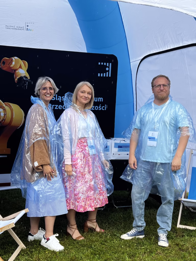 Zdjęcie przedstawia trzy osoby stojące pod niebieskim, dmuchanym namiotem podczas wydarzenia plenerowego. Wszyscy mają na sobie przezroczyste peleryny przeciwdeszczowe. Na pierwszym planie po lewej stronie stoi kobieta w okularach, z krótkimi blond włosami, ubrana w niebieską sukienkę w paski i białe tenisówki. Obok niej, w środku, stoi kobieta w różowej spódnicy i jasnej bluzce, również w pelerynie przeciwdeszczowej, z identyfikatorem na szyi. Po prawej stronie znajduje się mężczyzna z brodą i okularami, ubrany w jasnoniebieską koszulkę i dżinsy, także z identyfikatorem. W tle widać baner z napisem "Śląskie Centrum Przedsiębiorczości" oraz stoisko z materiałami promocyjnymi.