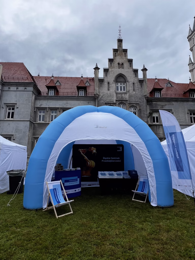 Zdjęcie przedstawia stoisko Śląskiego Centrum Przedsiębiorczości zorganizowane pod dużym, niebiesko-białym, dmuchanym namiotem na tle zabytkowego budynku. Namiot stoi na zielonym trawniku, a w jego wnętrzu znajdują się różne materiały promocyjne, takie jak broszury, ulotki oraz kilka opakowań. Na leżaku umieszczonym przed namiotem widnieje logo Funduszy Europejskich. Po prawej stronie namiotu widoczna jest biało-niebieska flaga z napisem "Fundusze Europejskie".