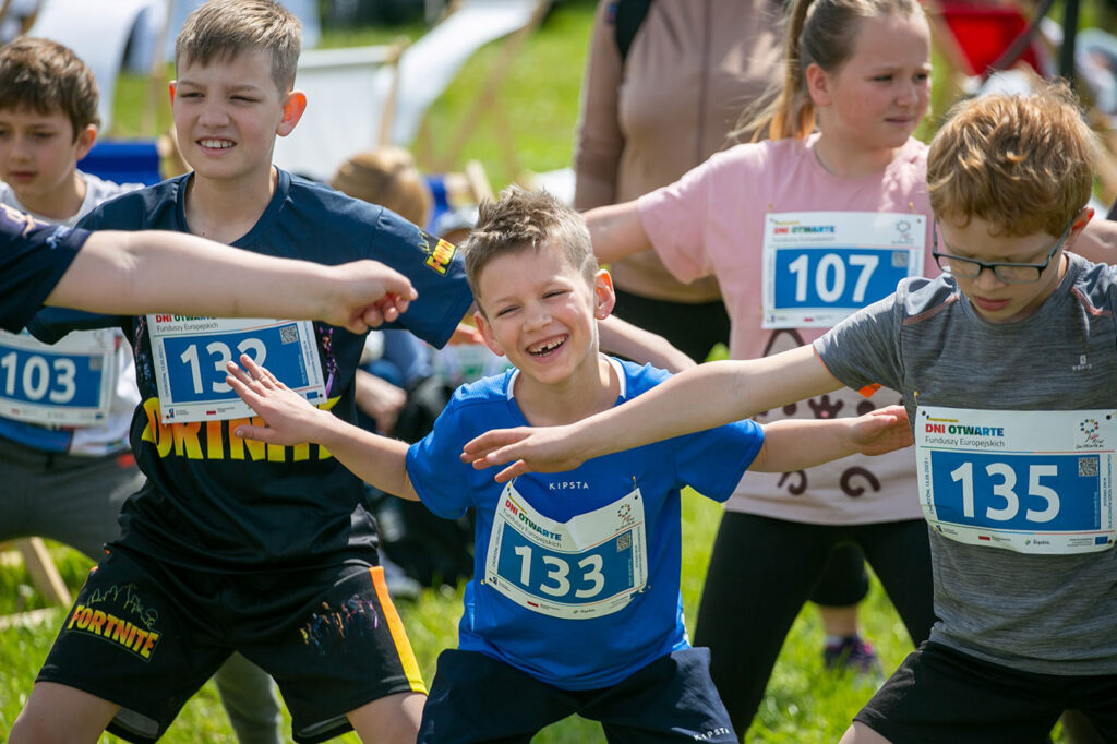 Na trawie ćwiczy grupa dzieci. Ubrane są w krótkie spodenki i koszulki z krótkimi rękawami w różnych kolorach, np. chabrowym, granatowym, szarym, niebieskim, różowym. Mają także plakietki z numerami startowymi, na których widoczne są napisy Dni Otwarte Funduszy Europejskich. Dzieci stoją w rozkroku i mają rozłożone ramiona. Chłopczyk w centrum kadru szeroko się uśmiecha, a pozostałe dzieci skupione są na ćwiczeniach.