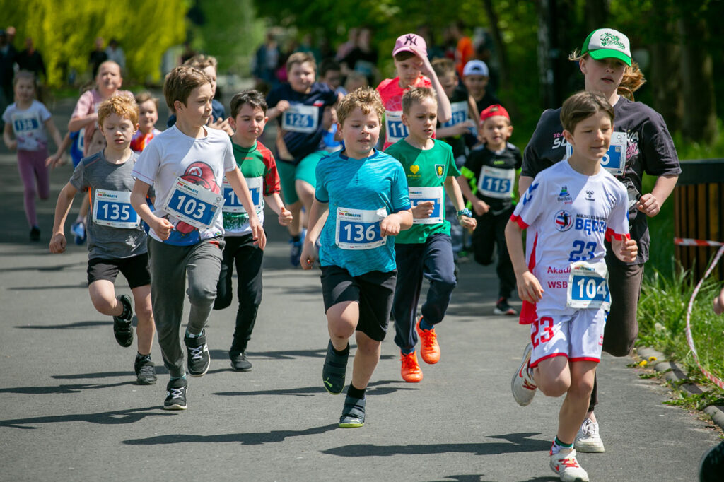 Duża grupa dzieci biegnie alejką. Na ich twarzach widoczne jest skupienie na zadaniu. W tle znajdują się drzewa i trawa, a także biało-czerwona taśma wyznaczająca trasę. Dzieci są ubrane w kolorowe koszulki, czapki i obuwie. Wszystkie mają plakietki z numerami startowymi, na których widoczne są napisy: Dni Otwarte Funduszy Europejskich.
