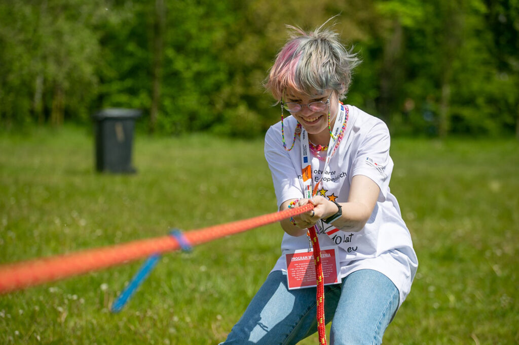Na tle łąki wśród drzew znajduje się młoda, roześmiana kobieta przeciągająca pomarańczowo-żółtą linę. Ma na sobie dżinsy oraz białą koszulkę z symbolem Dni Otwartych Funduszy Europejskich, a także okulary na łańcuszku składającym się z kolorowych kulek. Włosy ma pokryte farbami w pastelowych kolorach: jasnoszarym, łososiowym, różowym.