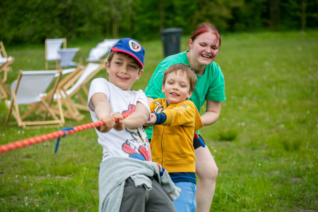 Na tle łąki i drzew pomarańczowo-żółtą linę przeciąga kobieta i dwóch chłopców. Wszyscy są uśmiechnięci. Mają na sobie kolorowe ubrania: zielone, żółte, niebieskie, białe. Za nimi po lewej stoją białe leżaki.