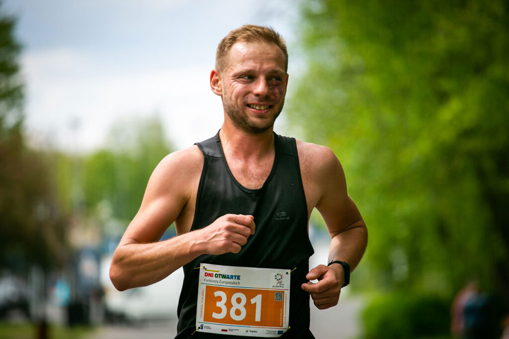 Na tle nieba i drzew widoczny jest zmęczony, ale uśmiechnięty biegacz w czarnej koszulce-bokserce, do której ma przypiętą plakietkę z numerem startowym i napisem: Dni Otwarte Funduszy Europejskich.