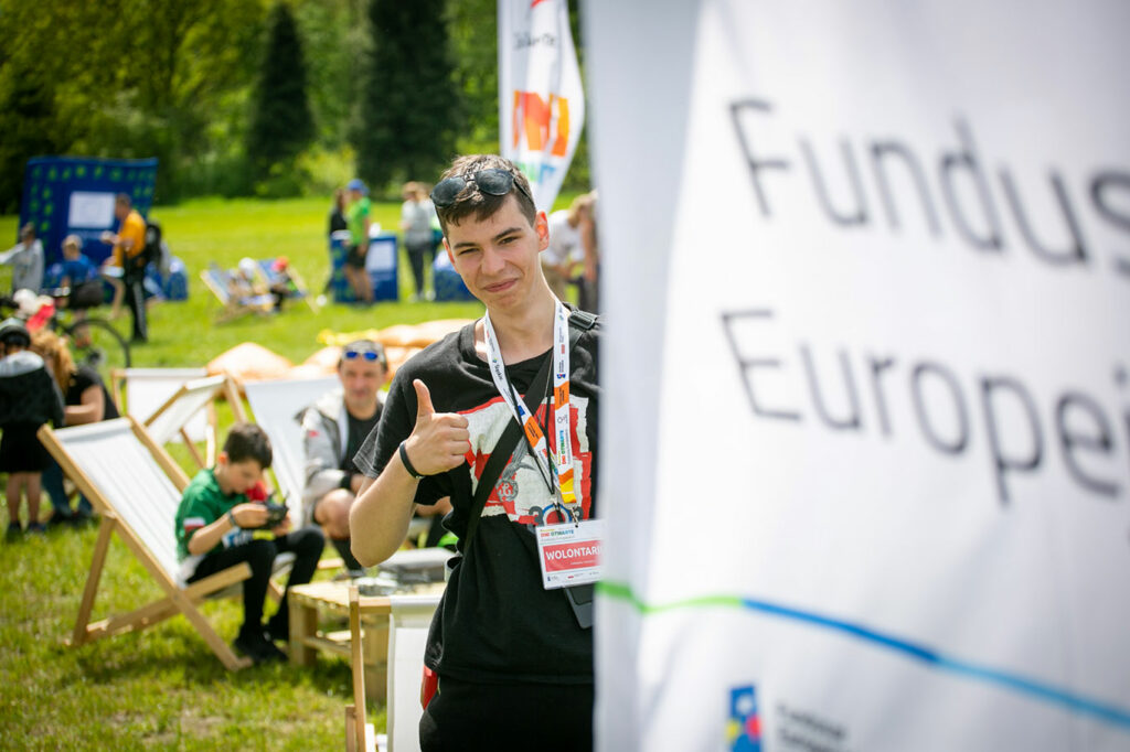 Ilustracją tekstu jest fotografia przedstawiająca młodego chłopaka z okularami przeciwsłonecznymi na głowie i plakietką „wolontariusz” zawieszoną na szyi, który pokazuje gest uniesionego w górę kciuka. Za nim znajdują się uczestnicy pikniku. Fotografia pochodzi z archiwum Urzędu Marszałkowskiego Województwa Śląskiego.