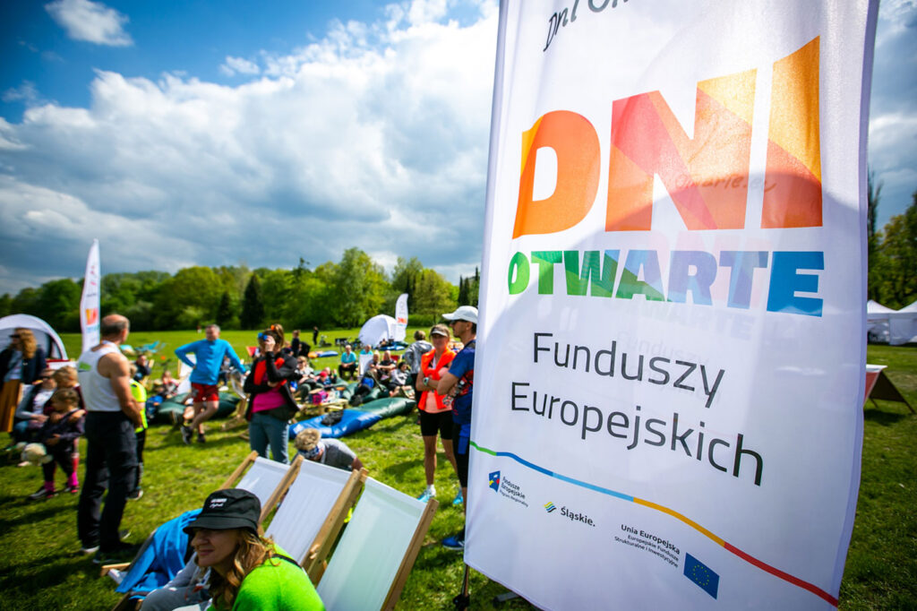 Na tle drzew i łąki znajduje się tłum kolorowo ubranych osób. Wszystkie w sportowych strojach. Niektóre stoją, inne siedzą na krzesłach, leżakach i workach. Niebo jest pogodne, błękitne z białymi chmurami. Na pierwszym planie znajduje winder z napisem: Dni otwarte Funduszy Europejskich.