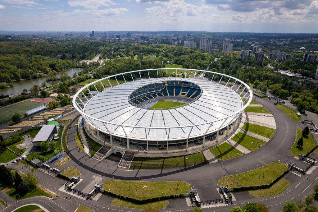 W centralnym miejscu znajduje się widoczna z lotu ptaka szara, okrągła bryła Stadionu Śląskiego wraz z otaczającymi go drogami i bramkami wejściowymi. W części wewnętrznej stadionu dominuje kolor niebieski i żółty. Widać także fragment murawy. W tle po prawej znajduje się kanał regatowy oraz zieleń Parku Śląskiego, a po lewej szare bloki Osiedla Tysiąclecia wraz z najwyższymi „kukurydzami”.