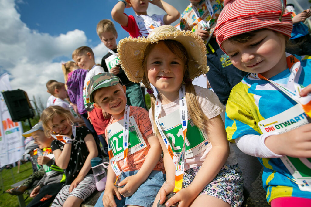 Na tle błękitnego nieba z białymi chmurami siedzi grupa kilkunastu uśmiechniętych dzieci, ubranych w kolorowe koszulki i krótkie spodenki. Dziewczynki mają rozwiane włosy. Jedna z nich ma na głowie dzianinową czapkę z serduszkiem, druga słomkowy kapelusz, a dwóch chłopców — czapki z daszkiem. Dzieci mają plakietki z numerami startowymi oraz medale na wstążkach, na których widoczne są napisy: Dni Otwarte Funduszy Europejskich.