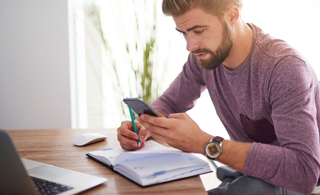 Fotografia przedstawia mężczyznę wypełniającego terminarz. W jednej ręce trzyma ołówek, którym coś zapisuje, w drugim zaś smartfon, z którego coś przepisuje.
