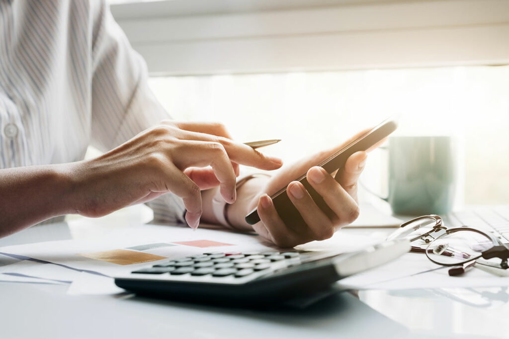 Na ilustracji widoczne są dłonie trzymające długopis i smartfona. Na blacie stołu leży również kalkulator.