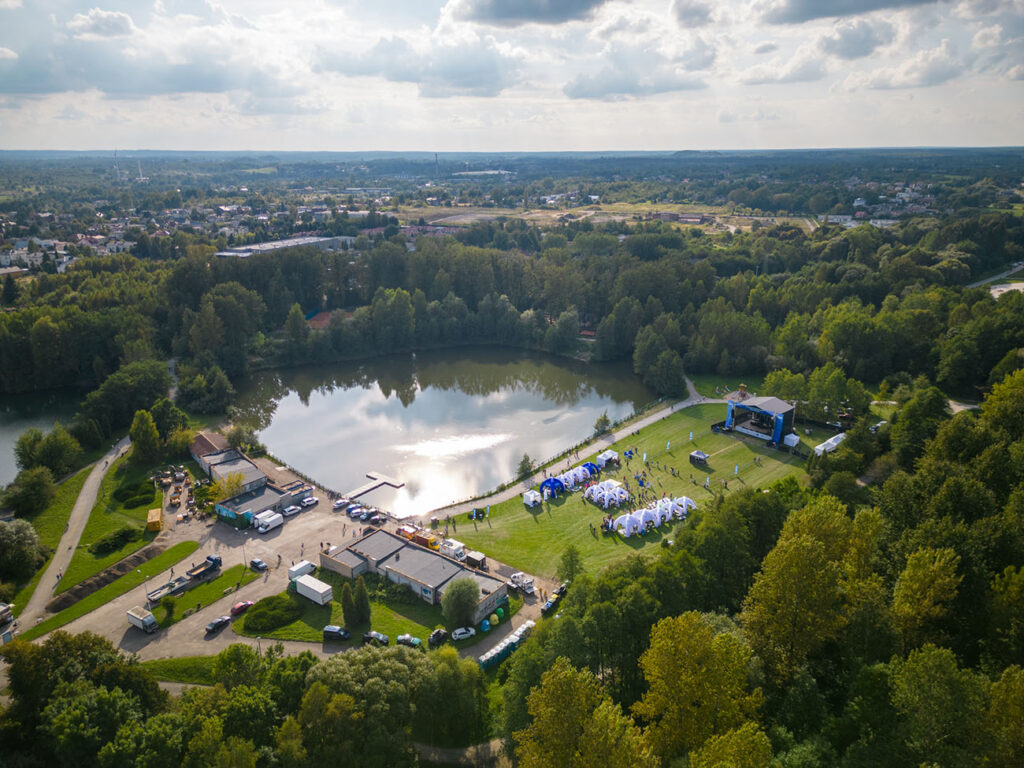 Ilustracja przedstawia park widziany z drona. Na środku jest duży staw, wokół którego znajduje się infrastruktura: budynki i namioty.