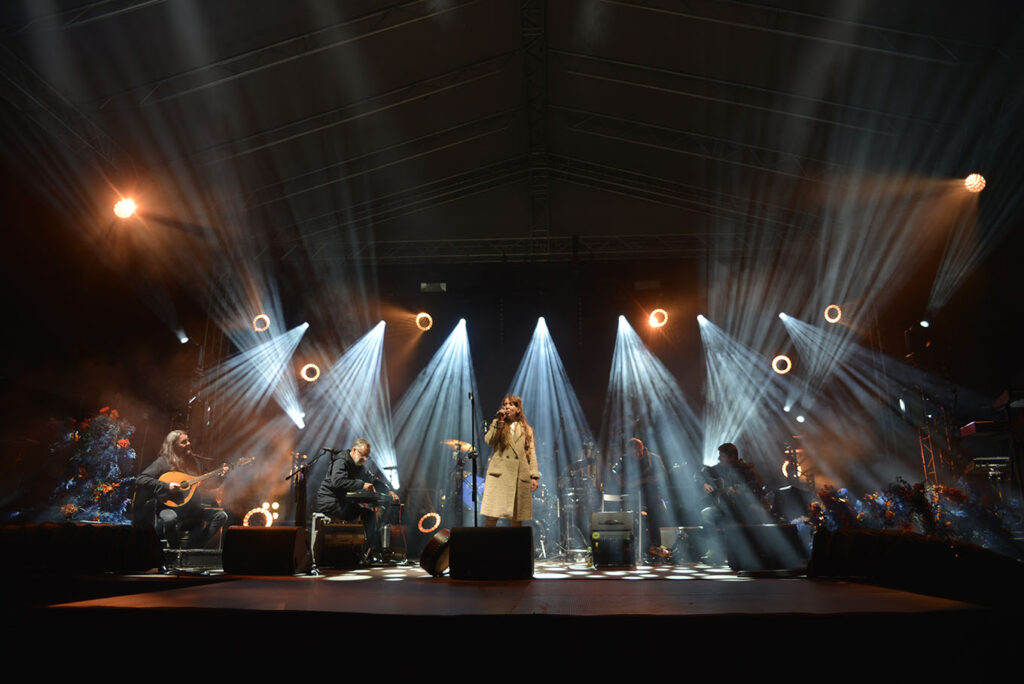 Na zdjęciu widać fragment koncertu. Na scenie stoi wokalista, z jej lewej i prawej strony siedzi zaś pięciu muzyków.