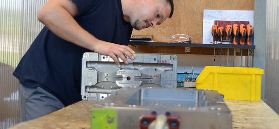 Zdjęcie przedstawia mężczyznę w czasie pracy w warsztacie. Za nim znajduje się panel, na którym zawieszone są śrubokręty i klucze.