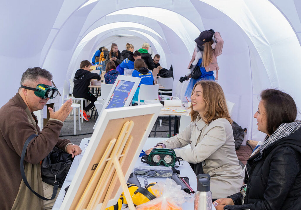 Zdjęcie wykonano we wnętrzu namiotu. Przy stolikach – stanowiskach, siedzi wiele osób. Na pierwszym planie widać mężczyznę w alkogoglach.
