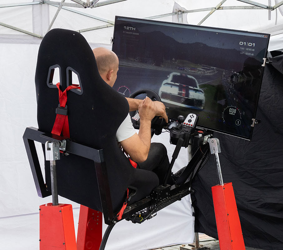 Na zdjęciu jest ukazany symulator jazdy samochodem. Składa się z ruchomej platformy na siłownikach, na którym zamontowane są fotel, monitor, kierownica, drążek skrzyni biegów i kilka różnych przycisków. Korzysta z niego mężczyzna.