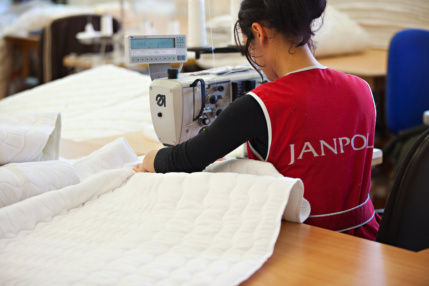 Zdjęcie przedstawia kobietę, która szyje na maszynie. Ma upięte włosy i ubrana jest w czerwony fartuch z napisem Janpol.