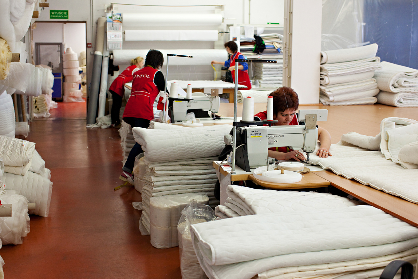 Fotografia przedstawia wnętrze szwalni. Na pierwszym planie, przy maszynie do szycia, siedzi kobieta. W tle widoczne są trzy inny kobiety, rozkładające materiał.