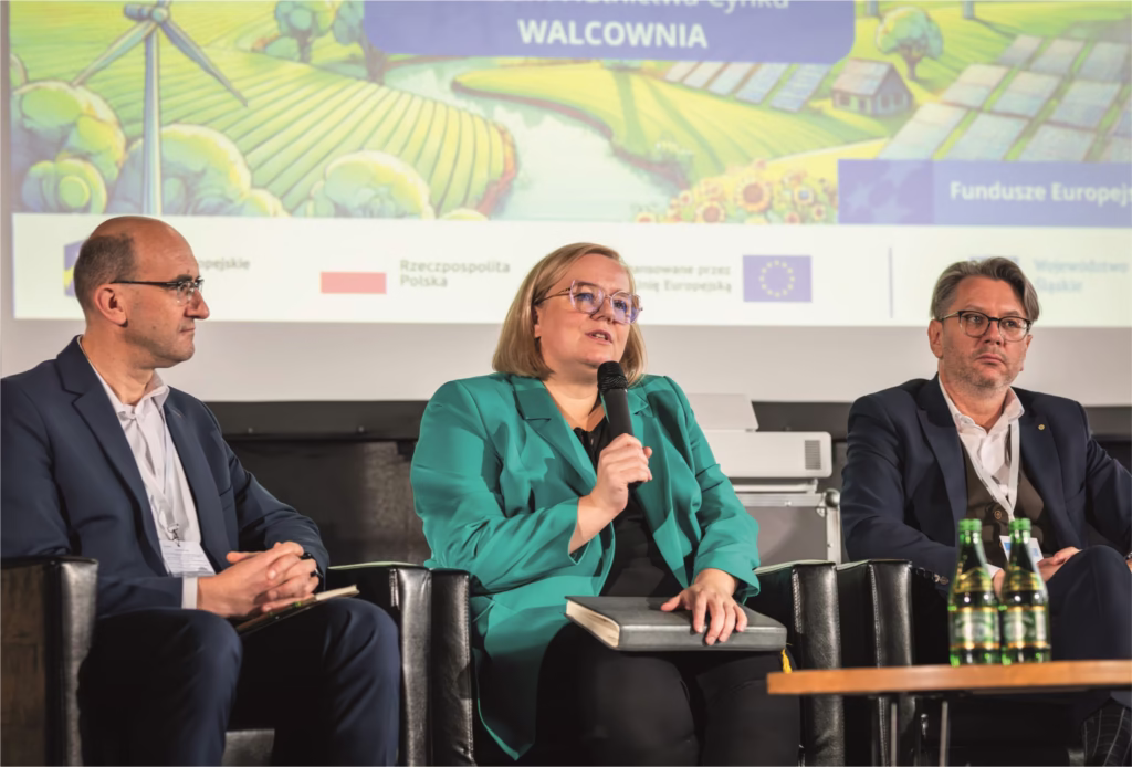 Na tym zdjęciu widzimy dwóch mężczyzn siedzących na scenie w czasie panelu dyskusyjnego a na środku kobietę z mikrofonem