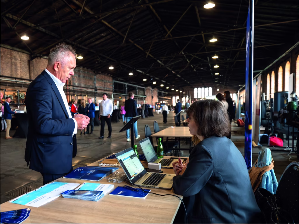 Na zdjęciu widzimy kilka osób przy recepcji konferencji. Mężczyzna w szarym garniturze podpisuje dokument na wysokim, niebieskim stole z napisem "Fundusze Europejskie dla Śląskiego". Obok niego stoją dwie kobiety oraz młody mężczyzna, którzy prawdopodobnie obsługują recepcję.