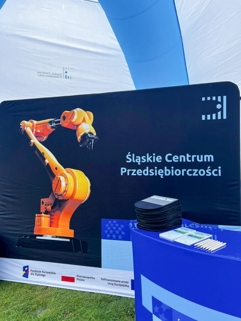 W namiocie ustawiona jest ścianka ze zdjęciem ramienia maszyny CNC i napisem: Śląskie Centrum Przedsiębiorczości. Na pierwszym planie znajduje się trybunka, na której leżą gadżety reklamowe.