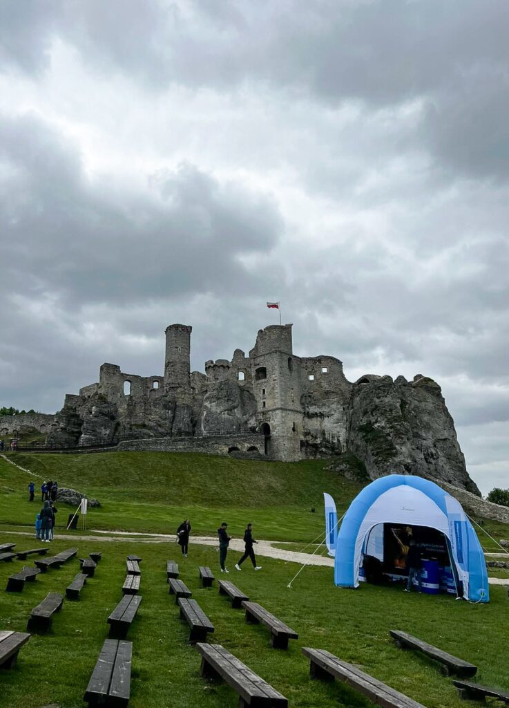 W pochmurny dzień u stóp zamku rozstawiono namiot z symbolami Funduszy Europejskich. Wokół namiotu i na drodze do zamku widoczni są ludzie.