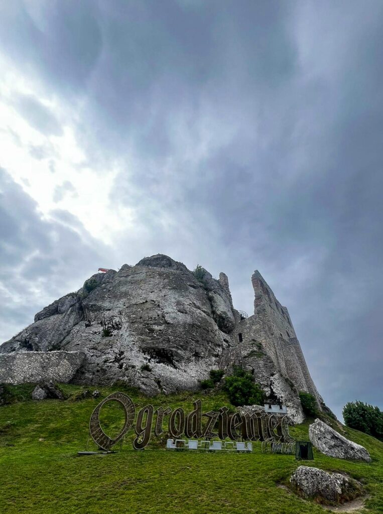 Na tle skał i ruin zamku widoczny jest napis: Ogrodzieniec. Niebo zasnute jest chmurami.