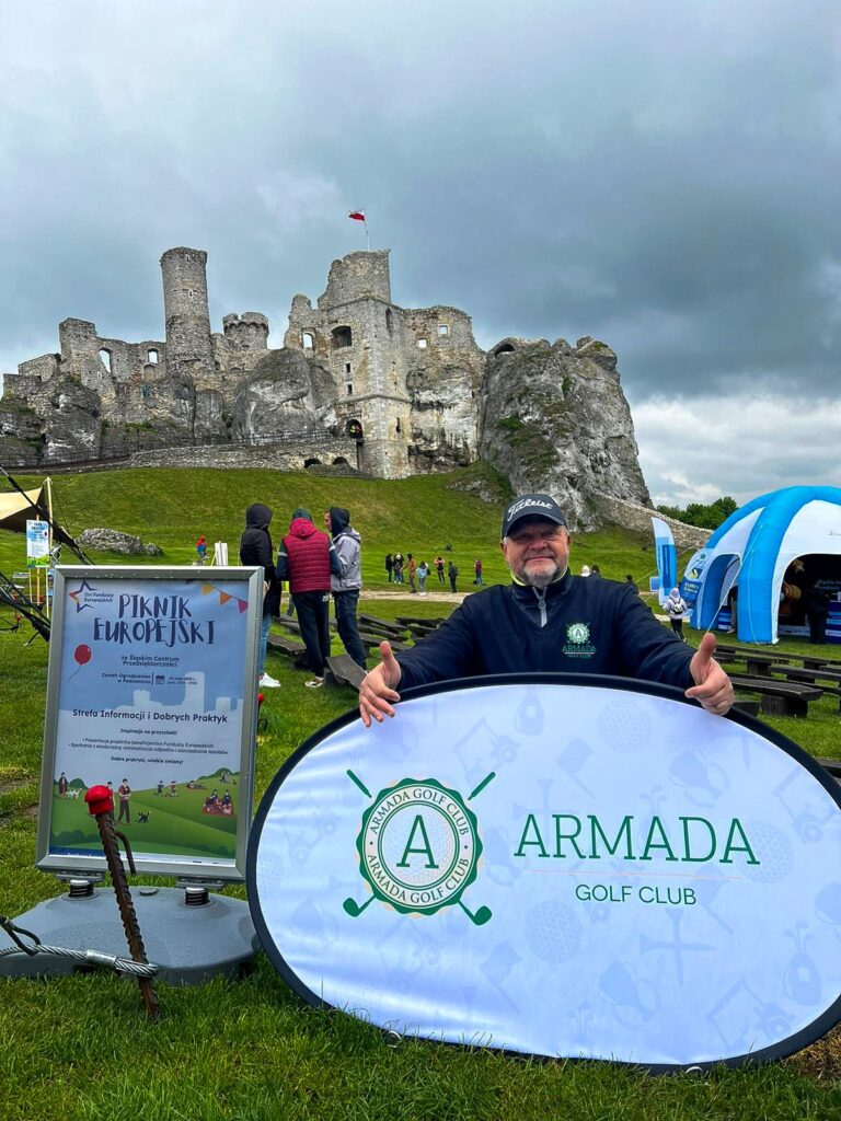 Uśmiechnięty mężczyzna w czapce z daszkiem prezentuje baner Armada Golf Club. Obok stoi plakat Pikniku Europejskiego z napisem Strefa Informacji i Dobrych Praktyk. W tle widoczni są uczestnicy pikniku, namiot oraz Zamek Ogrodzieniec.