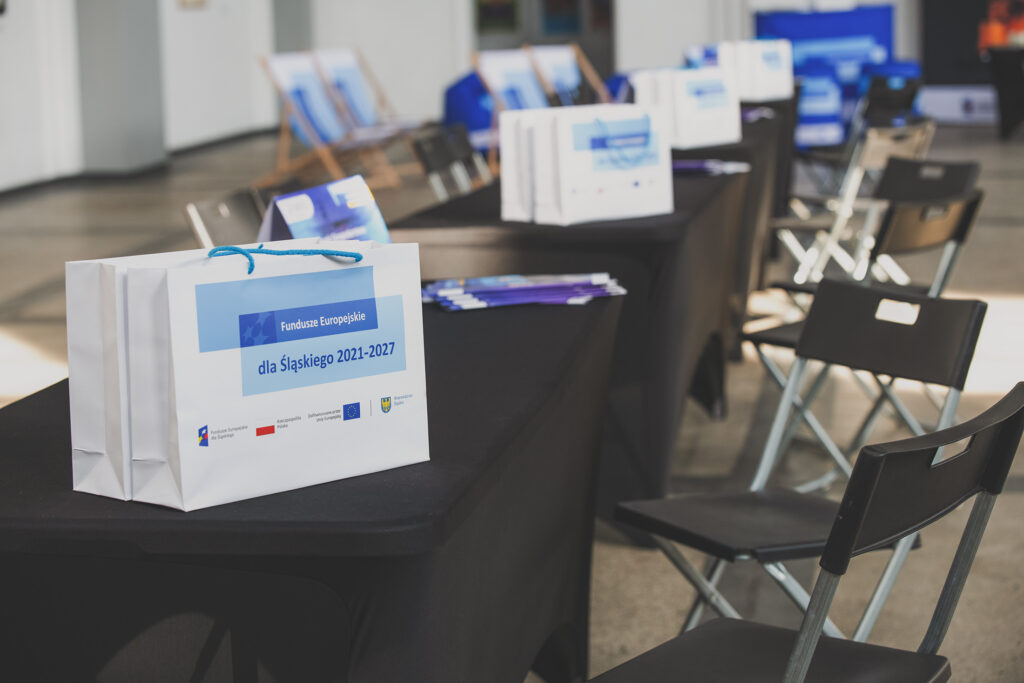 Zdjęcie przedstawia rząd krzeseł oraz stolików przykrytych czarnymi tkaninami. Na stolikach widoczne są torby z motywem przewodnim Funduszy Europejskich, napisem Fundusze Europejskie dla Śląskiego 2021-2027 oraz logotypami.