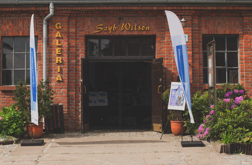 Zdjęcie przedstawia wejście do budynku, na którym znajduje się napis: Galeria Szyb Wilson. Przed budynkiem widoczne są krzewy kwitnących rododendronów, flagi z motywem przewodnim Funduszy Europejskich oraz plakat z napisem Czas na zmiany.