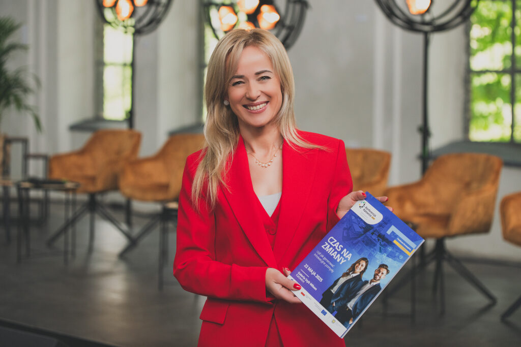 Na zdjęciu widoczna jest uśmiechnięta blondynka w czerwonym żakiecie i kamizelce oraz białej bluzce. W dłoniach trzyma folder, na którego okładce przedstawieni są kobieta i mężczyzna w biznesowych, granatowych strojach. Obok nich znajduje się napis: Czas na zmiany, czyli jak generować innowacyjne pomysły, 22 maja 2025 Galeria Szyb Wilson Katowice. Na górze folderu znajduje się motyw przewodni Funduszy Europejskich. W tle za stojącą kobietą umieszczony jest rząd brązowych foteli oraz lamp.