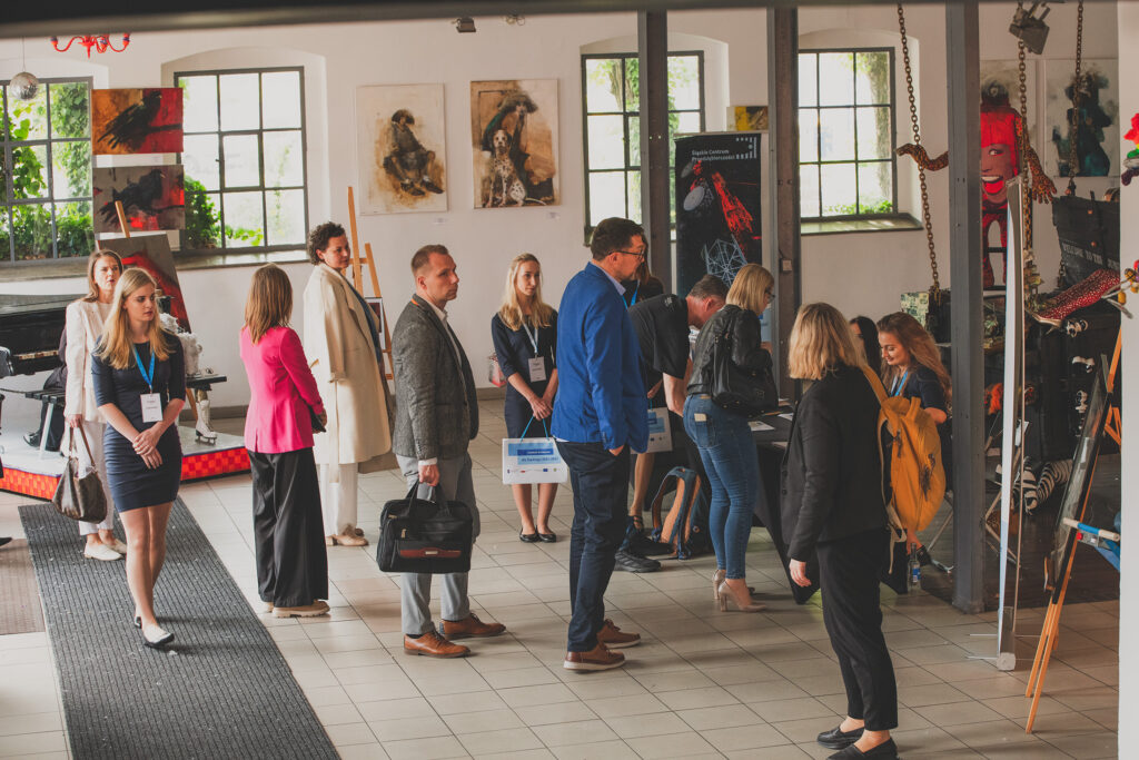 Zdjęcie przedstawia wnętrze wypełnione obrazami oraz instalacjami. Przed stolikiem, za którym siedzą hostessy, ustawiła się kolejka osób.