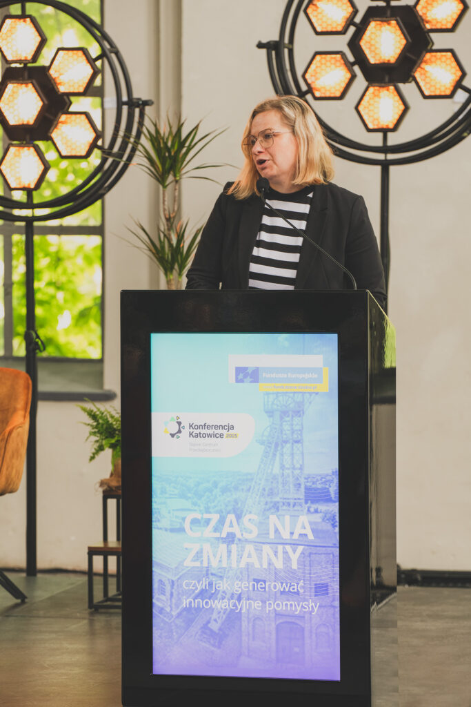 Za pulpitem stoi kobieta ubrana w pasiastą biało-czarną bluzkę oraz czarny żakiet. Na przedzie mównicy widoczne jest zdjęcie z szybem kopalnianym i napisem: Czas na zmiany, czyli jak generować innowacyjne pomysły. Powyżej napisu znajduje się motyw przewodni Funduszy Europejskich oraz logo z zębatym kołem i napisem Konferencja Katowice.