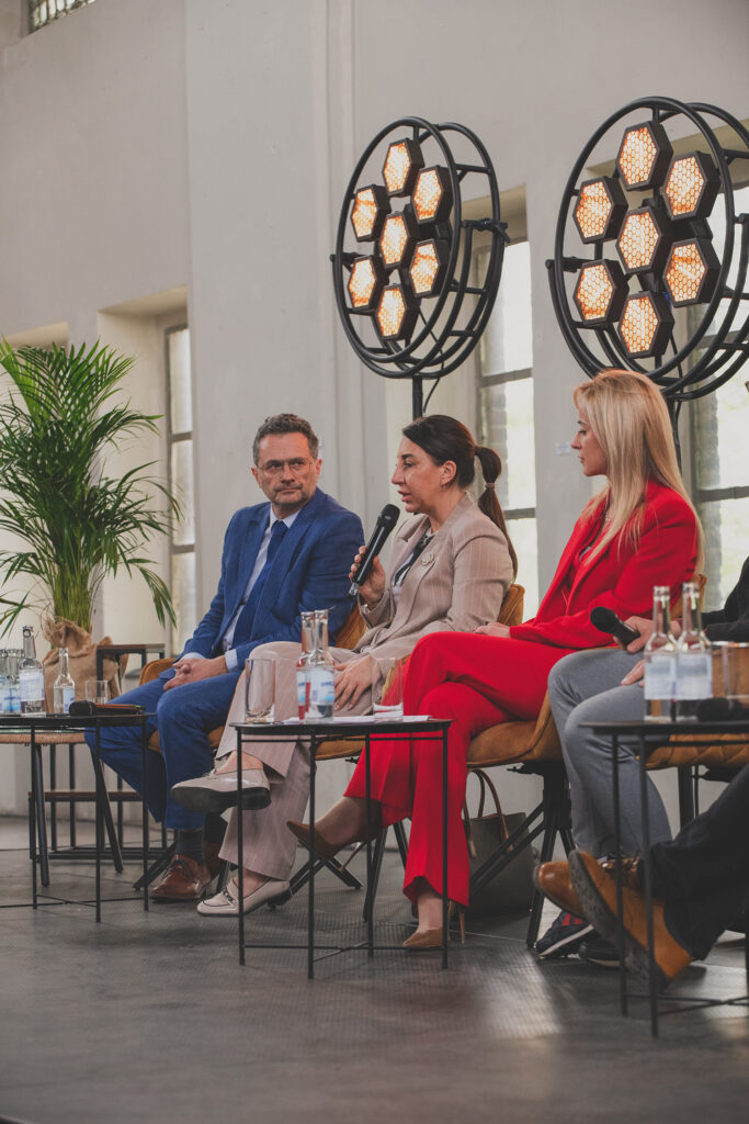 Na zdjęciu widoczni są prelegenci - dwie kobiety i mężczyzna. Wszyscy ubrani w biznesowe stroje. Jedna z kobiet trzyma w dłoni mikrofon. W tle widoczne są lampy okna oraz okazała palma.