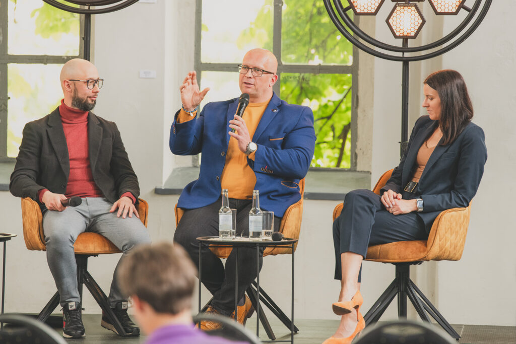 Zdjęcie przedstawia gestykulującego prelegenta z mikrofonem w dłoni. Po jego jednej stronie siedzi kobieta, po drugiej mężczyzna. W tle widoczne są okna, a na pierwszym planie głowa osoby siedzącej na widowni.