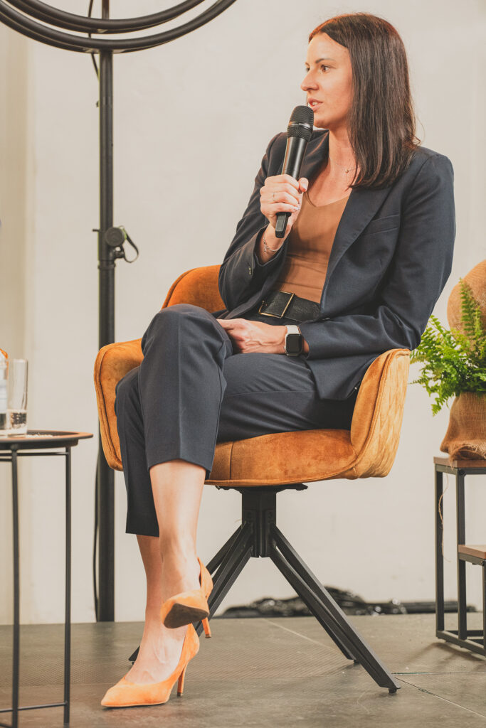 W brązowym fotelu siedzi kobieta trzymająca w dłoni mikrofon.